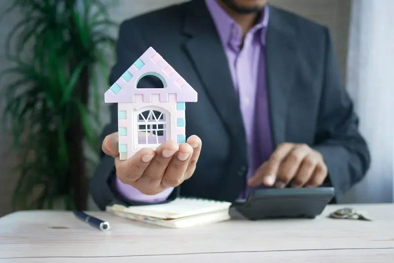 A man holding a small model house next to a calculator